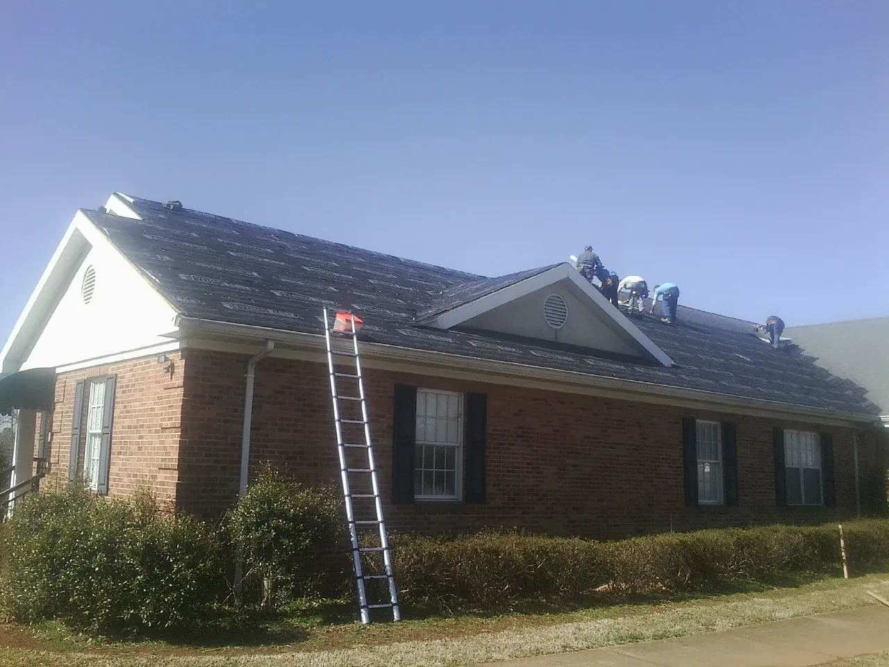 Crew tearing shingles off a brick ranch roof with a ladder set — Emergency Roof Repair work by our crew in Rutland